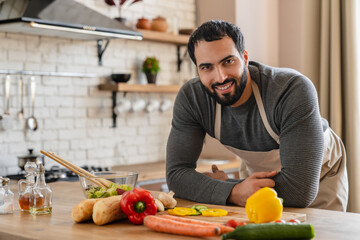 A portrait of a cheerful adult bearded Arabian man in apron , leaning on a kitchen counter and preparing a vegetable salad while looking at camera and smiling