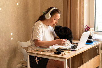 Woman Embroidering While Wearing Headphones at Home Desk. Senior black cat sits beside her, while a...