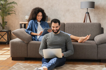 Couple sitting on living room sofa using digital technology devices while working remotely from...