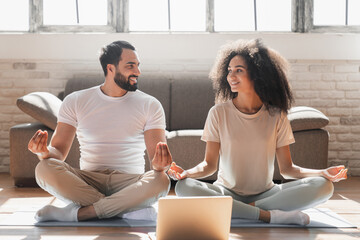 Calm mixed race couple in pajamas meditating, listening spiritual practices lessons on laptop, sitting on lotus pose looking to each others at home light living room . Yoga concept.