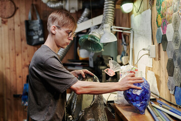 Young male worker of plastic recycling workshop taking packed bottle caps and putting them into large cellophane sack