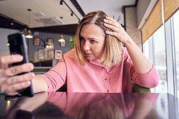 Woman in pink shirt checking hair with phone mirror in modern cafe setting.