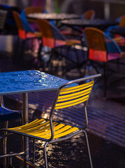 An outdoor cafe with colorful chairs, wet tables, and dramatic lighting.