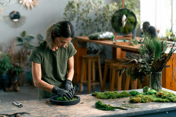 in large green workshop a thin woman with green hair in black gloves at a large table collects a...