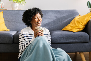 girl sitting in living room with cup of hot coffee enjoying under blanket. Joyful black girl...