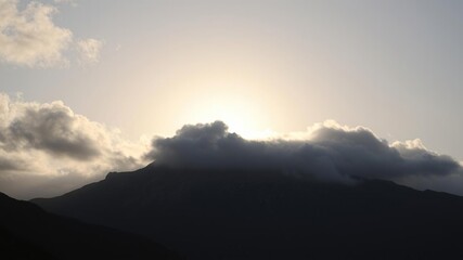 A majestic sun is hidden behind thick grey clouds, casting a dramatic shadow over a rugged mountain range, rocky, peaks