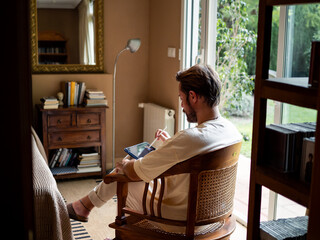 A man sits comfortably in a wooden chair, engrossed in working on his tablet. The room features...