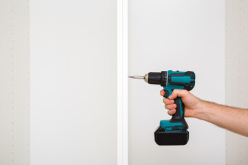 Young adult man hand using professional battery screwdriver and screwing screw in white board of base cabinet frame. Assembling new home wooden furniture. Closeup. Front view.