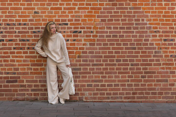 Full length front view portrait of beautiful serene long haired caucasian blond woman in white sweater with bag standing outdoor on city street by red brick building wall. Copy space. Fashion theme.