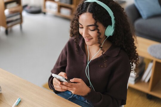 A woman with curly hair and green headphones smiles as she looks down at her smartphone, seated in a relaxed indoor environment, creating a friendly and approachable atmosphere.