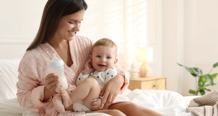 Mother holding cute little baby and bottle of milk on bed at home. Space for text