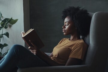 A black woman in casual attire, reading a book with focus and serenity. She appears relaxed in her own home, engaging in a quiet moment of leisure.