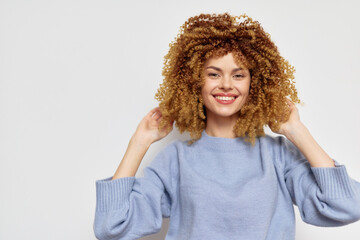 Happy young woman with curly hair, smiling and posing against a light grey background, wearing a cozy sweater, showcasing a cheerful and vibrant personality