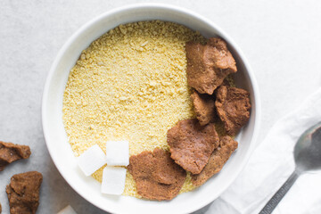 Overhead view of kuli-kuli and yellow garri in a white bowl, top view of nigerian groundnut cakes and garri in a bowl, fresh homemade nigerian kulikuli and cassava in a ceramic bowl