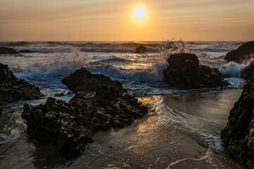 large, rugged rocks rise from the ocean, framing a setting sun that casts a warm, golden light over the waves