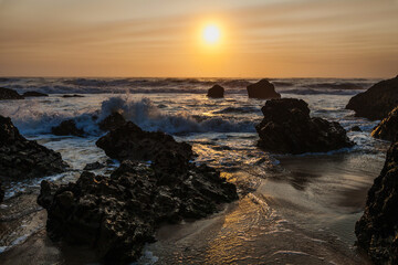 coastal sunset scene, waves rush in from the open sea, filling the space between the dark, rugged rocks