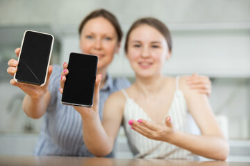Happy mother and teenage daughter showing together their cellphones sitting at kitchen-table