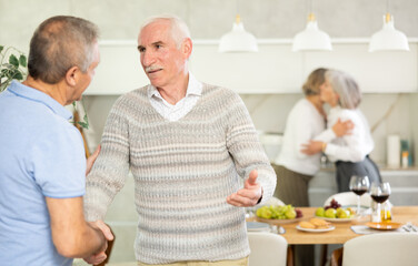 In cozy homely atmosphere, elderly male friends shake hands, say goodbye after socializing and gatherings. Their girlfriend is in background.