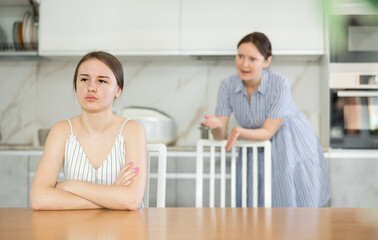 Upset daughter is sitting at table. Mom yells at her daughter in the background