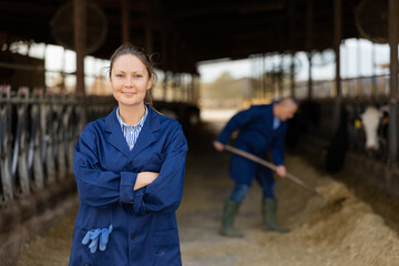 Portrait of woman ranch worker standing in cowshed near rows of cows in stalls with arms crossed.