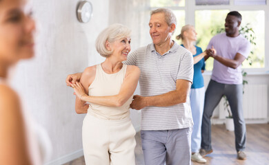 Obraz premium Senior man learns to dance Latin salsa dance with elderly lady. Dance studio mature clients move in background