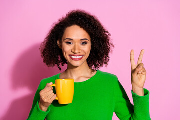 Photo portrait of attractive young woman hold coffee mug show v-sign dressed stylish green clothes isolated on pink color background