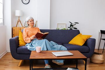 Senior woman wrapped in a warm blanket reading a book relaxing on sofa at home. Leisure and domestic lifestyle concept.