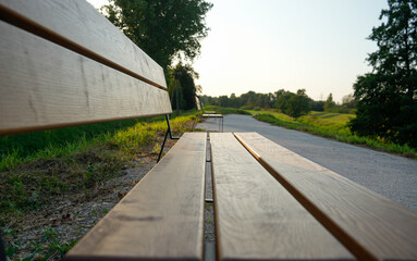 Close-up of a wooden bench along a path in a quiet park. The tranquil setting with green grass and trees is bathed in soft late evening light, relax and unwind in nature