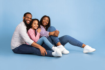 Cool Team. Beautiful and happy smiling young black family hugging and having fun time together while sitting on the floor and looking at camera, isolated on blue studio background, free copy space