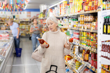 Elderly woman choosing hot chili sauce for herself in supermarket