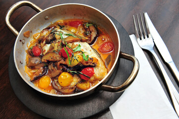 Top view of cooked Mediterranean Maghreb spicy fish served in a metal fry pan on restaurant table with knife fork and napkin.