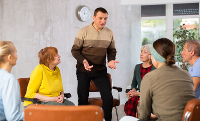 Portrait of positive adult man leading education class for group of elderly people, discussing...