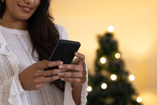 Asian woman texting on smartphone near Christmas tree, enjoying holiday season, at home - Powered by Adobe