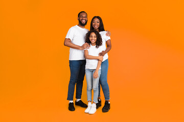 Full body length of excited African American man, woman and girl standing isolated on yellow orange studio wall, vertical shot. Smiling black husband and wife posing with daughter looking at camera