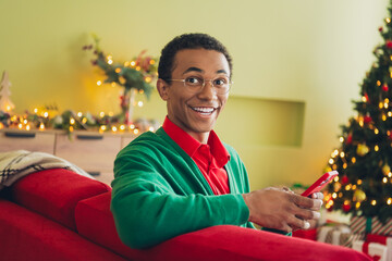 Photo of nice young man sit couch hold smart phone wear green cardigan christmas festive time...