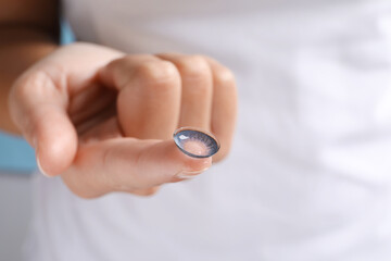 Woman holding color contact lens on light blue background, closeup