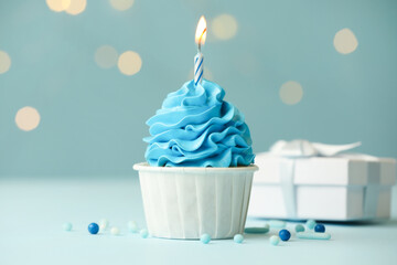 Birthday cupcake with burning candle and gift on light blue table against blurred lights