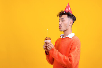 Portrait of Young Asian Guy Wear Birthday Cap Blowing Candle on Cupcake With Copy Space on Yellow Background