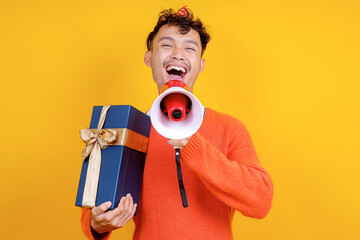 Happy Young Asian Man Shouting Using Megaphone While Holding Gift Box Isolated on Yellow Background