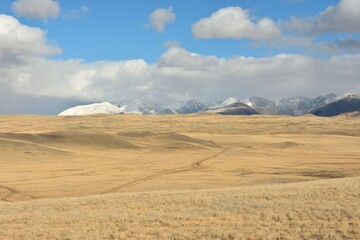 Hilly bare steppe with a network of roads under a cloudy sky against the backdrop of snow-capped mountains on an autumn sunny day.
