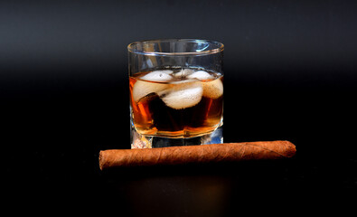 A glass of whiskey with ice and a large Cuban cigar on a black background.