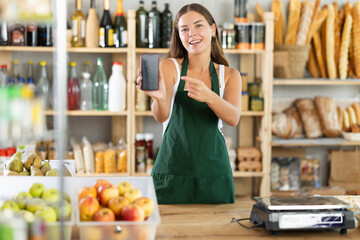 Young female seller in uniform showing information on smartphone screen in grocery store