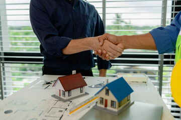engineer and architect are shaking hands to celebrate because they just finished big construction project, two engineers do handshake after the meeting, blueprint is on the desk