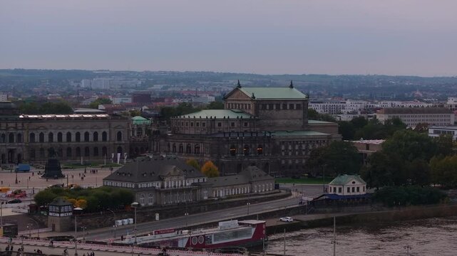 Tourists stroll near the Semperoper Opera House in Dresden, Germany, as the Elbe River flows alongside the iconic building during twilight, creating a picturesque urban scene