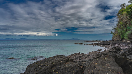 The coast of a tropical island at low tide. Exposed boulders of the water's edge. The turquoise ocean is calm. Green vegetation on a coastal hill. Mountains on the horizon. Blue sky, clouds.Madagascar