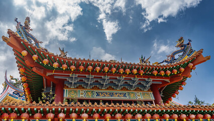 The roof of a Chinese temple against a blue sky, clouds. Rows of red traditional lanterns suspended along the curved edges. Ornaments. Colorful figures of dragons, birds on the roof ridge. Malaysia.