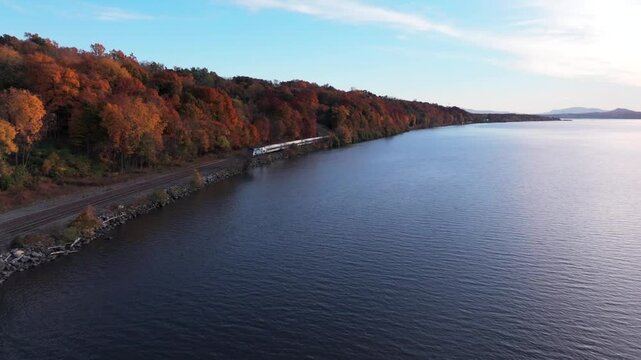 An aerial view over the Hudson River next to train tracks on a sunny day in autumn. The camera dolly in towards an outbound Amtrak train moving fast ahead in the distance.
