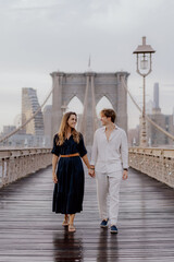 A couple strolls hand in hand on Brooklyn Bridge, surrounded by a stunning skyline and beautiful...
