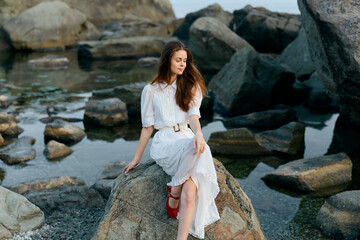 Serene woman in white dress on ocean rock gazes at horizon, red shoes add vibrant contrast