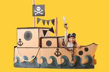Cute little girl dressed as pirate playing with sword in cardboard ship on yellow background © Pixel-Shot